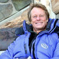 Man in blue snowsuit sitting against wall and smiling to camera.